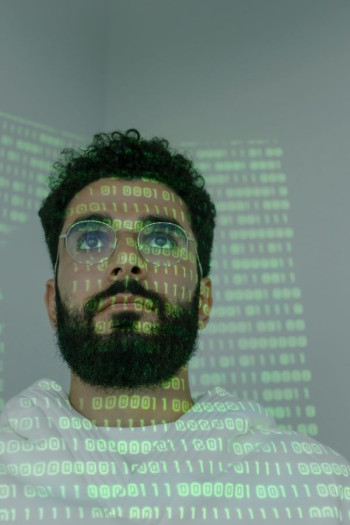 A close up image of a man's head and shoulders. He has dark hair and a beard as well as glasses. He is wearing a hoody. He has a projection over his face and body of computer code.