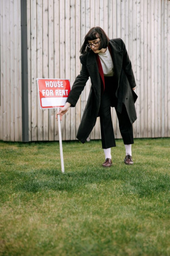 A white woman with a dark bob haircut pokes a 'for rent' red sign into grass. She is outside a house. She is wearing glasses and a dark suit.