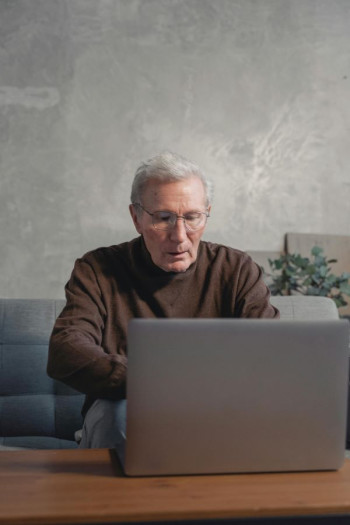 An older white man in a brown high-necked top sits on a couch looking at an unbranded, open laptop.