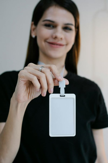 A white woman with long dark straight hair and wearing a black t-shirt holds out a blank identification badge.