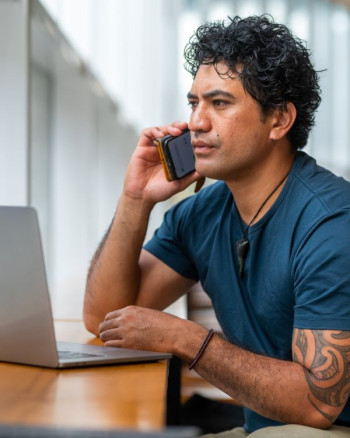 A Māori man with black curly hair and an arm tattoo is talking on a cellphone. He wears a blue t-shirt and has a laptop open in front of him.