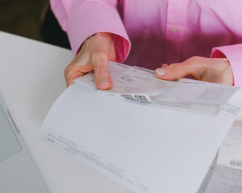 A white woman's hands hold receipts and a piece of paper that we assume is a bank statement. She is wearing a pink shirt.