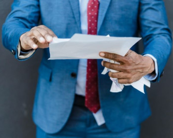 A man in a blue suit with red tie is cropped so you can see his torso and arms. He is dark skinned. He is holding papers.