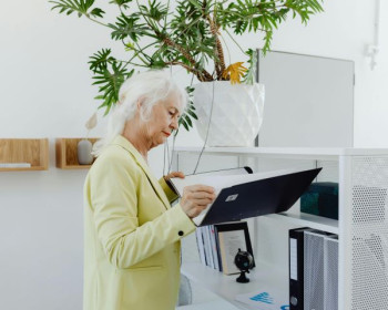 An older white woman with white hair and a light yellow blazer looks at a file in an office.