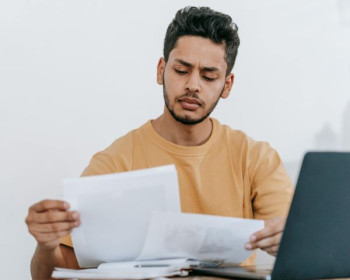 A man with dark hair and beard looks at some paper documents. He seems to be concentrating.