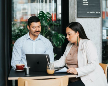 A man and a woman sit at a cafe table looking at a laptop. The man has dark hair and a moustache and is wearing a light blue shirt. The woman has long, dark, straight hair and a moko on her chin. She is wearing a beige blazer and a brown top. 