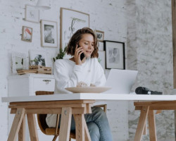 A woman in a white top sits at a trestle table desk talking on her smartphone.