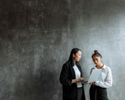 Two woman dressed in black and white business clothes stand talking about something that is shown on a laptop that one is holding. Their stance is formal; a professional conversation. They stand in front of a contemporary polished concrete wall. 