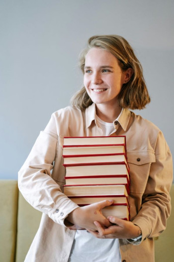 A white woman with blonde shoulder length hair holds a stack of eight hardcover books. She is wearing a white shirt and beige jacket. We can't see her legs. She looks happy.