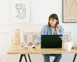 A woman in her 20s or 30s sits at a wooden trestle table in a contemporary home office setting. She is looking at a notepad on the desk and has an open laptop in front of her. She is white with a brown bob haircut and is wearing a casual blue shirt.