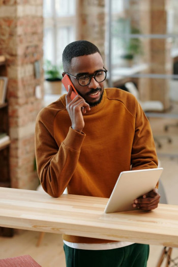 A black man in a pumpkin spice coloured jersey talks on a red smartphone while looking at a tablet. He is leaning on a wooden high table. He has glasses.