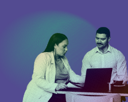 A man and a woman sit at a table looking at an open laptop. 