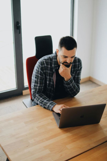 A man with dark hair and beard looks at a laptop on a table. He is wearing a checked shirt and has his chin in his hand.