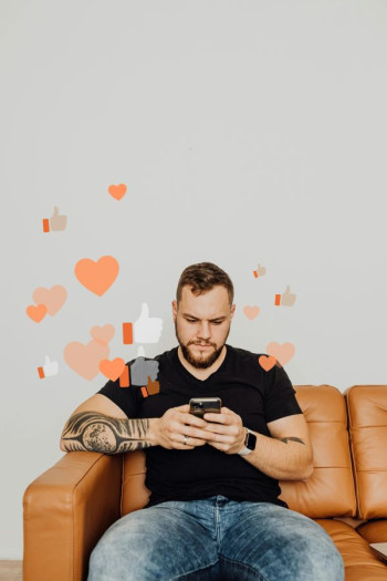 A white man with arm tattoos sits on a light brown couch. He is wearing blue jeans and a black t-shirt. He has brown hair and a beard and is looking at a mobile phone that has graphics of hearts and 'like thumbs' coming out of it. 