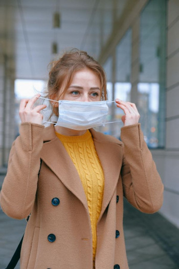 A white woman puts a white medical mask on. She has strawberry blonde hair that is tied up. She is wearing a yellow jersey and a tan coat.