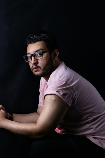 A man in a pink t-shirt sits side on, slightly forward with his elbows on his knees. His short sleeve is rolled up to show a plaster on his bicpep. He has dark hair, facial hair, and glasses.