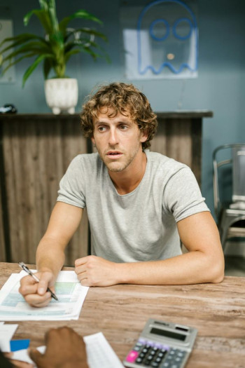 A man with light brown curly hair sits at a desk filling in a form. A calculator is on the table. He wears a short sleeved plain grey tshirt.