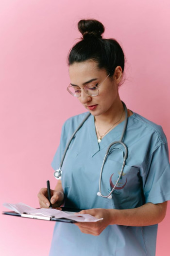 A woman in a blue scrubs top writes with a pen on a clipboard with documents that she is holding. She has dark hair and glasses. The background is a plain pink wall.