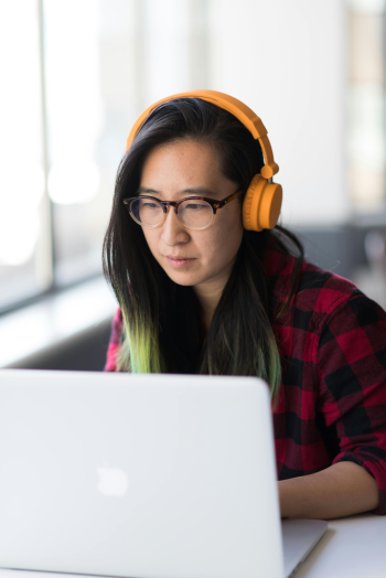 A woman with long dark hair that has green ends looks at a laptop. She is wearing glasses and orange headphones.