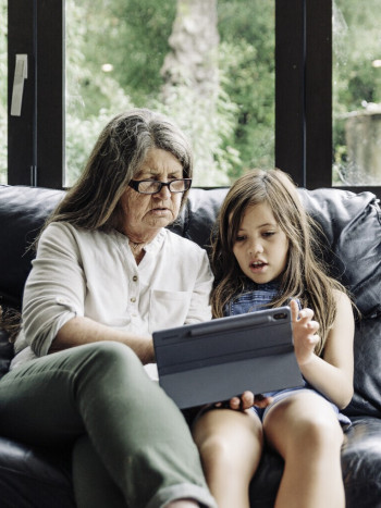 An older woman wtih long grey hair and glasses sits on a black couch with a child of about 10. They are looking at a tablet together. The woman wears khaki trousers and a cream shirt. The child is wearing denim overalls.