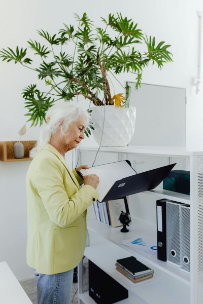 An older woman with white hair wearing a yellow blazer and jeans looks at files in a lever arch file.