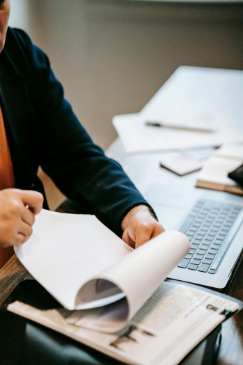 A persons arms on a desk showing a laptop and papers. The person is reviewing a document.