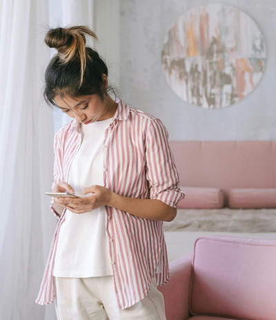 A woman with a top-knot looking at a phone in her hand. She wears beige pants, white top, and striped shirt which is open.