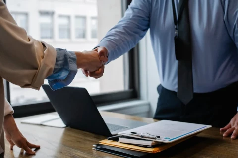 Two people shaking hands over a desk with documents on it.