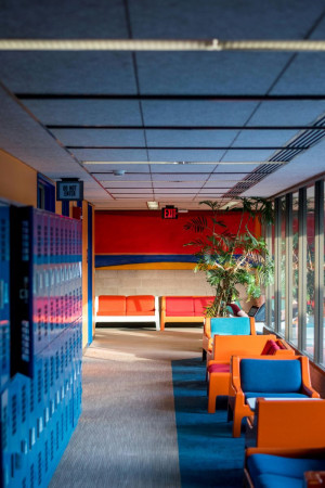 A scene of a school inspired lounge. Blue lockers line the left wall. There is a blue run and the chairs and sofas are all orange and blue.