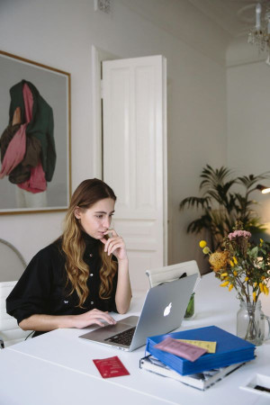 A white woman with long curly brown hair looks at a laptop on a table. Her finger is at her mouth, wondering. She is in a contemporary environment with flowers on the table.