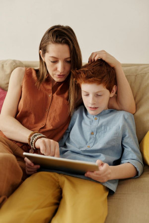 An adult woman and a boy of about 11 sit on a soft couch looking at an iPad or other tablet. The woman’s hand is on the boy’s head and they’re both looking at the screen.