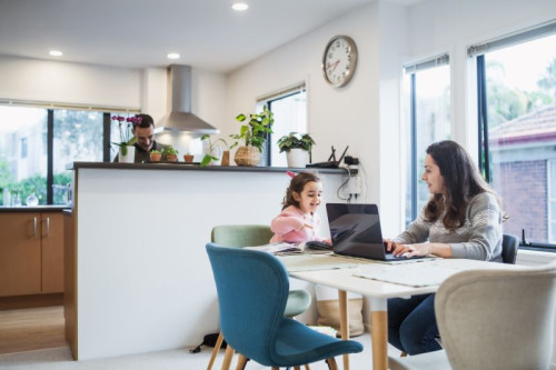 A woman with brown hair sits in front of her open laptop at the kitchen table. A child is beside her engaging with what the woman is doing.