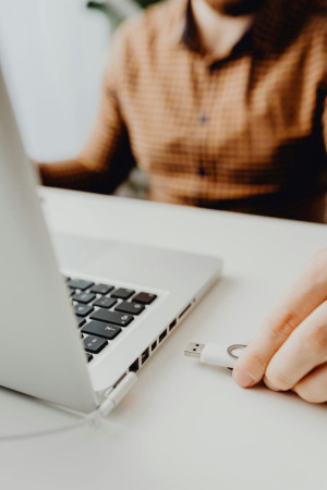 A cropped image of a laptop open on a desk. A person's hand is about to put a USB drive into a port.