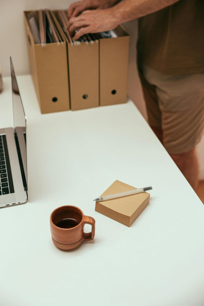 Three brown filing boxes are sitting on a white table with a person's hands rifling through them. In the foreground there is a cube of beige post it notes and a cup of coffee.