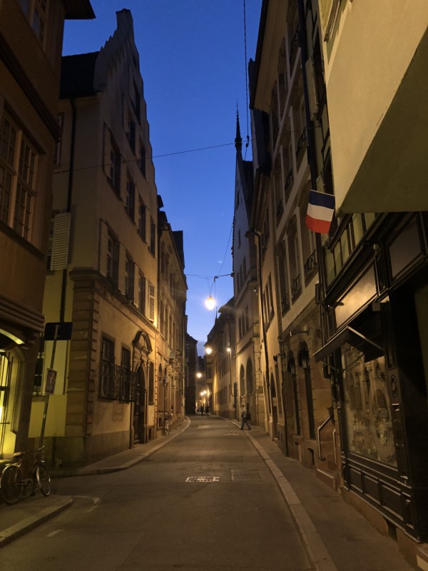A dark street lined with stone buildings. A french flag hangs from one.